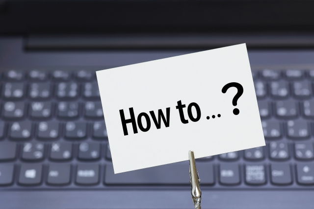 ノートパソコンのキーボード上に「How to…？」と書かれた紙とペンを置いたイメージ